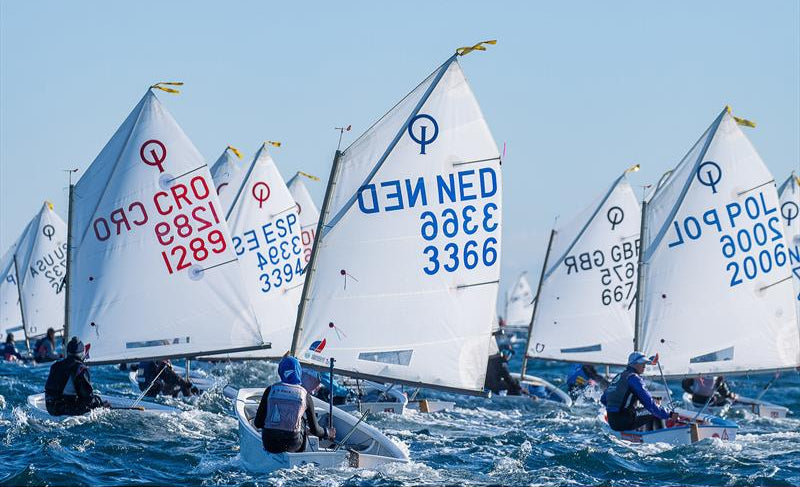 Optimist-class boats racing under the sun, part of a major youth sailing regatta—highlighting the foundations of foilboarding and marine sport progression.